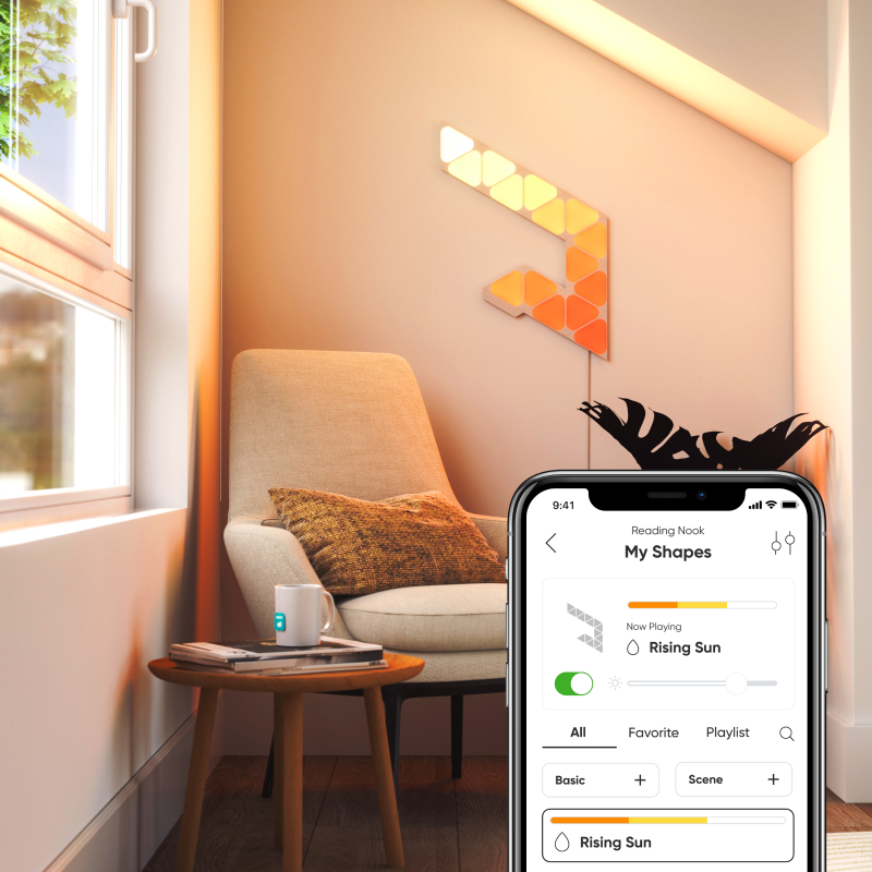Cozy reading nook with 9 Nanoleaf Shapes Mini Triangle light panels mounted on the wall beside a chair and a iPhone with the Nanoleaf Smarter Series mobile app on the screen.